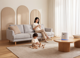 Woman sits on the gray sofa holding a mug, while a child sits on the floor playing with wooden blocks. Carepod One Plus humidifier rests on a wooden coffee table, next to a white bowl filled with apples.