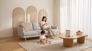 Woman sits on the gray sofa holding a mug, while a child sits on the floor playing with wooden blocks. Carepod Cube humidifier rests on a wooden coffee table, next to a white bowl filled with apples.