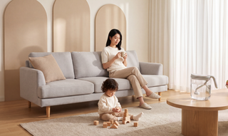 Woman sits on the gray sofa holding a mug, while a child sits on the floor playing with wooden blocks. Carepod Cube humidifier rests on a wooden coffee table, next to a white bowl filled with apples.