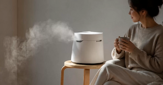 White humidifier emitting mist on a wooden stool next to a person holding a mug